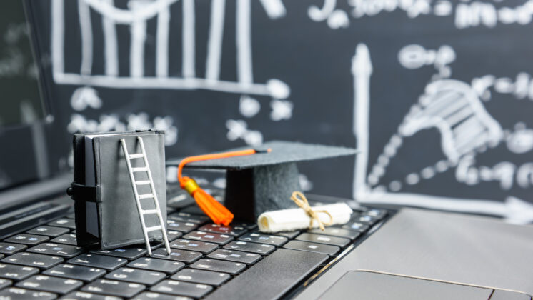 Graduation hat and miniature ladder sit on a keyboard with MBA graphs in the background to show digital entrepreneurs educate online