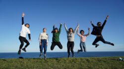 Six young people jump together on a grassy field with the ocean in the background