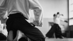Black and white image of aikido student kneeling with blurred partners training in the background