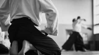 Japanese Gods, Art, and Management Theories in the Modern Era Black and white image of aikido student kneeling with blurred partners training in the background