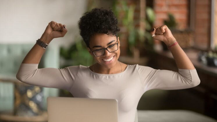 A Black woman in glasses raises her arms in triumph in front of a computer.