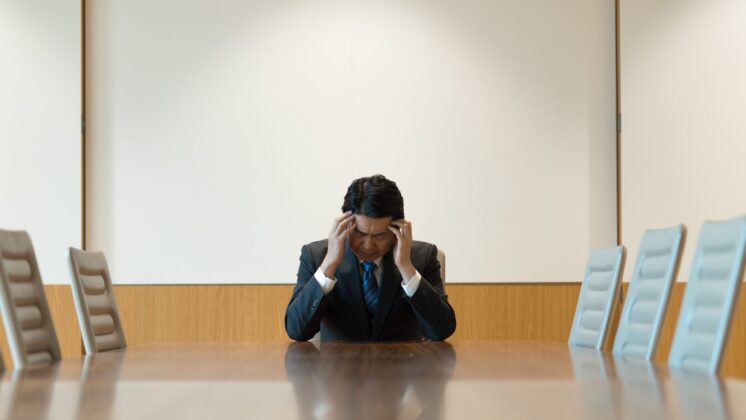 Businessman at an empty boardroom table holds his head in his hands in shame over bankruptcy