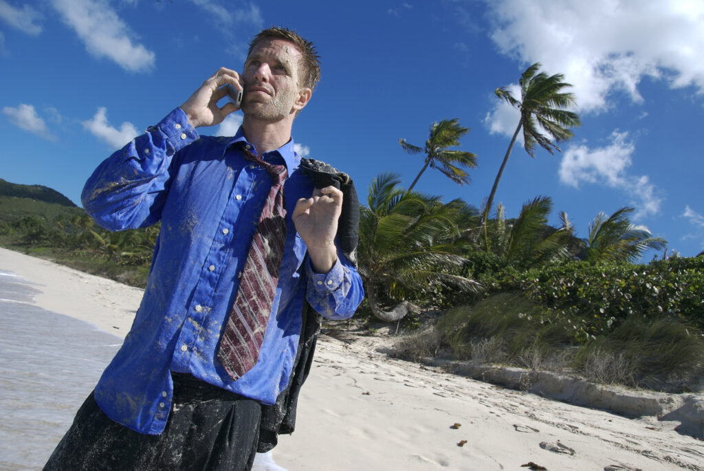 Businessman in disheveled clothing tries to find cell reception on a desert island