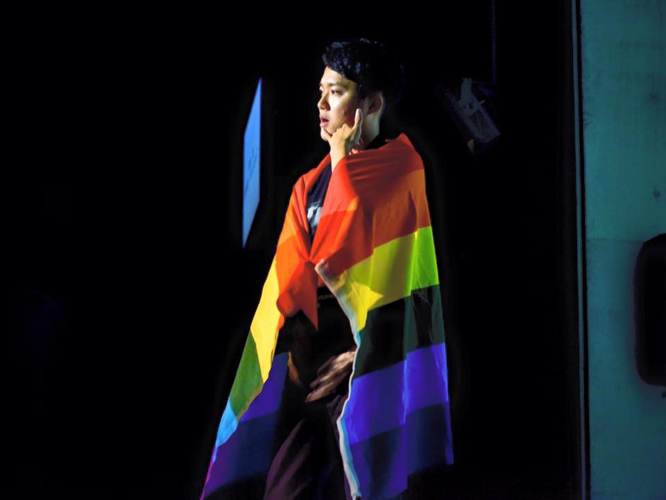 Image of Kan speaking while wrapped in a rainbow flag