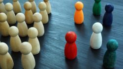 Unpainted pawns stand in strict rows facing colorful, dispersed pawns on a table, representing a workforce failing to integrate true DEI