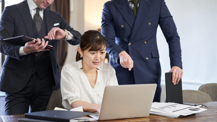 Japanese woman works in a panic; two bosses stand behind her with body language for poor psychological safety in the workplace
