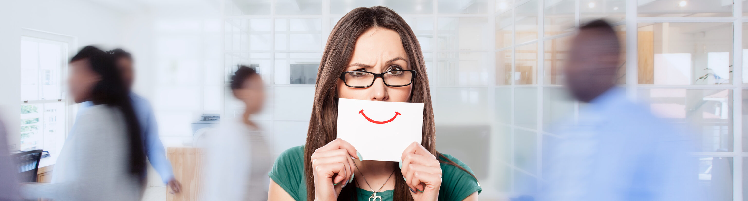 A distressed woman in an office with toxic positivity holds a smiley face on a card over her mouth