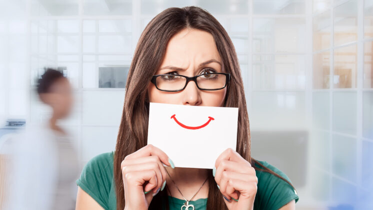 A distressed woman in an office with toxic positivity holds a smiley face on a card over her mouth