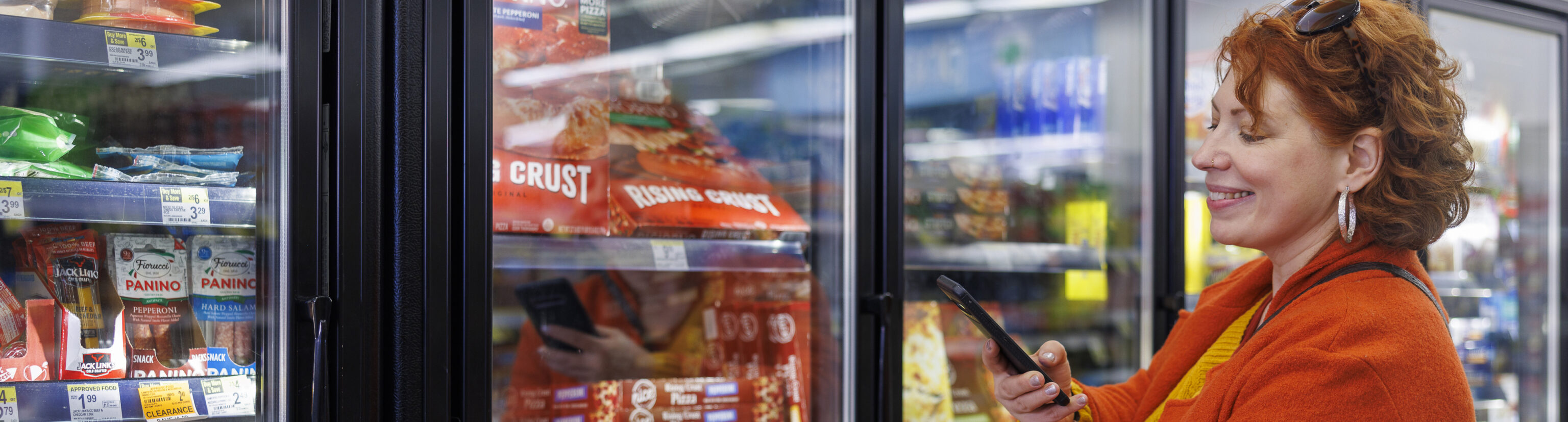 Smiling mature Caucasian woman in bright orange coat, using her smartphone to scan the label of a frozen food item in supermarket freezer section.