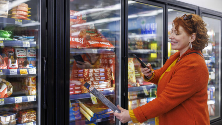 Smiling mature Caucasian woman in bright orange coat, using her smartphone to scan the label of a frozen food item in supermarket freezer section.