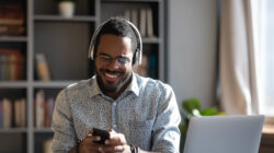 What’s the deal with the attention economy and how can we regain control? African man worker or student sit at desk distracted from study or work hold smartphone wear headphones hearing voice audio message from friend, enjoy free time use modern gadget wireless tech concept