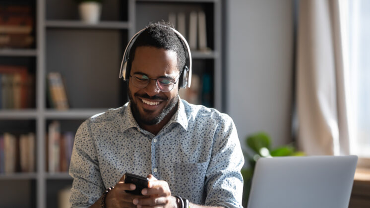 African man worker or student sit at desk distracted from study or work hold smartphone wear headphones hearing voice audio message from friend, enjoy free time use modern gadget wireless tech concept